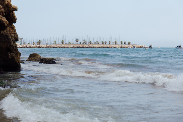 Камені і морські хвилі на узбережжі з пірсом і човнами на задньому плані в Каталонії (Іспанія).  - Фото, зображення