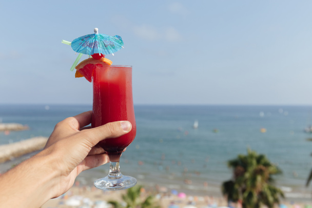 Обрізаний вид людини, що тримає склянку коктейлю з морським пейзажем та блакитним небом на фоні
  - Фото, зображення
