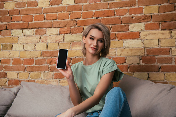 pretty woman smiling at camera while sitting on sofa and showing smartphone with blank screen - Фото, изображение