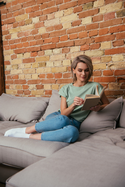 beautiful young woman reading book while sitting on sofa near brick wall - Photo, Image