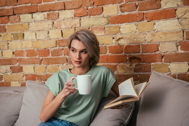 zamyślona młoda kobieta trzyma filiżankę herbaty i otwartą książkę siedząc na kanapie w pobliżu cegły ściany - Zdjęcie, obraz
