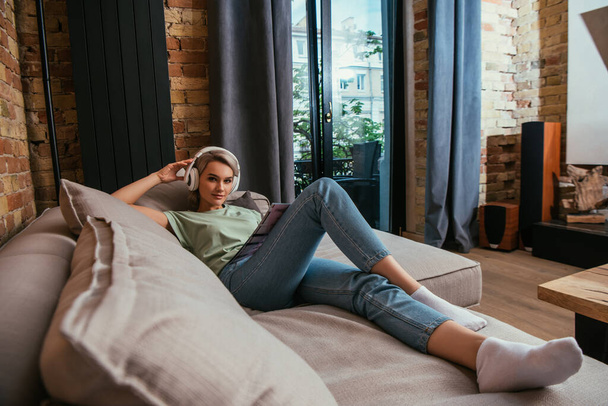 selective focus of beautiful woman in wireless headphones looking at camera while chilling on sofa  - Foto, Imagem