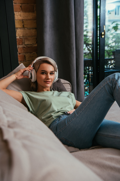 selective focus of attractive woman in wireless headphones smiling at camera while relaxing on sofa - 写真・画像