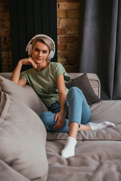 beautiful, dreamy woman in wireless headphones sitting on sofa and looking at camera - Foto, Imagem