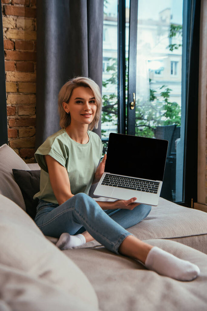 atractiva joven mujer sonriendo a la cámara mientras se sienta en el sofá y muestra el ordenador portátil con pantalla en blanco
 - Foto, Imagen