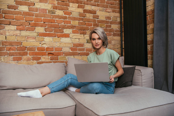 beautiful young woman looking at camera while sitting on sofa with laptop - Foto, Bild