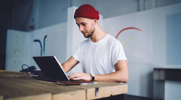 Conteúdo trabalhador adulto hipster casual profissional em vermelho beany e t-shirt sentado na mesa de madeira olhando para a tela do notebook no escritório moderno
 - Foto, Imagem