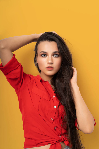 Cheerful mixed race brunette in red blouse isolated on yellow background. Beauty surprised woman with long dark hair and clean skin looking at camera while touching her hair with hands - Foto, afbeelding