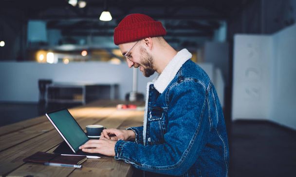 Side view of happy smiling casual adult worker in red beany and jeans jacket sitting at wooden desk typing at notebook at modern office - Foto, Bild