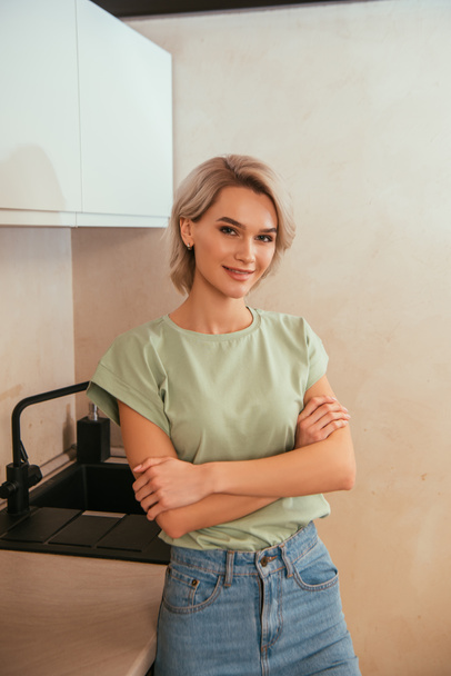 hymyilevä, kaunis nainen seisoo ristissä kädet ja katselee kameraa keittiössä - Valokuva, kuva