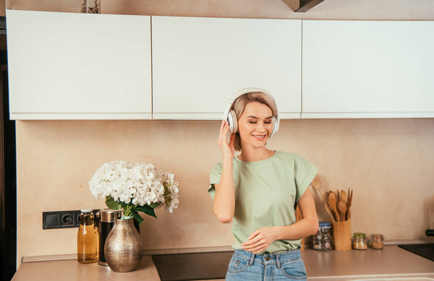 happy young woman listening music in wireless headphones in kitchen with closed eyes - Фото, зображення