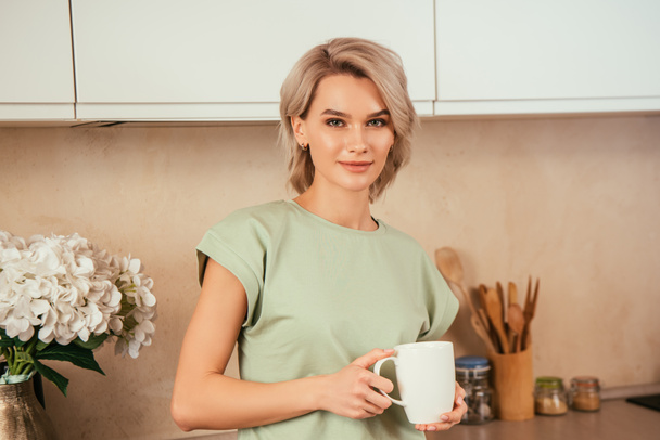 αυτοπεποίθηση, ελκυστική γυναίκα κοιτάζοντας κάμερα, ενώ κρατώντας ψηλά το τσάι στην κουζίνα - Φωτογραφία, εικόνα