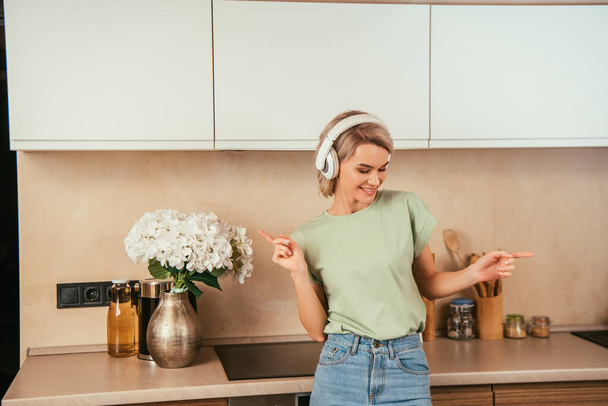 happy young woman gesturing while listening music in wireless headphones in kitchen - Zdjęcie, obraz