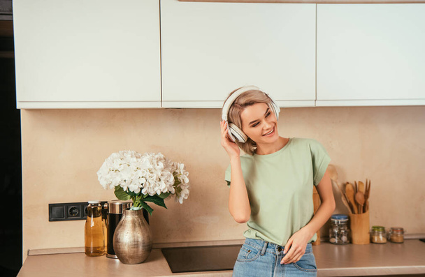 smiling, attractive woman listening music in wireless headphones in kitchen - Фото, изображение