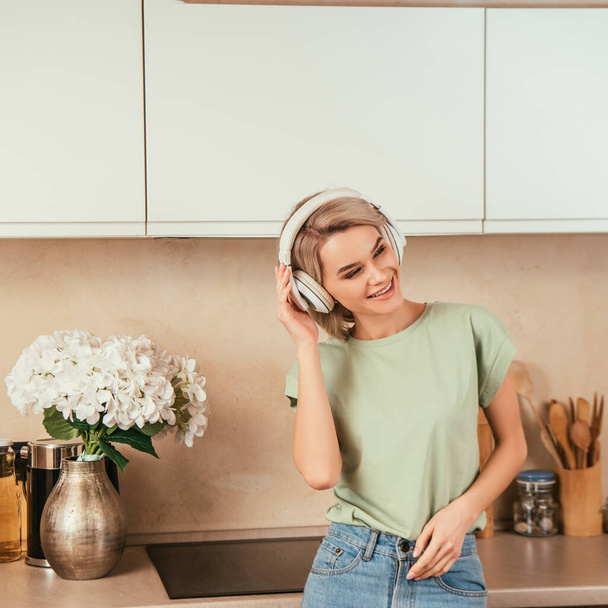 szczęśliwa młoda kobieta słuchanie muzyki w bezprzewodowych słuchawkach w kuchni - Zdjęcie, obraz