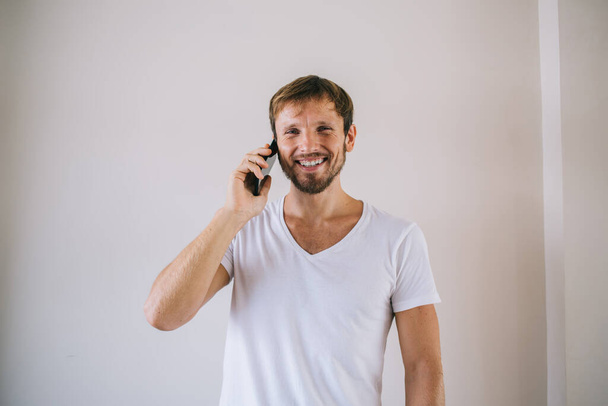Happy Caucasian male model in white t-shirt with copy space area for your brand name or label posing for camera while making positive smartphone cell call via application for communication indoors - Foto, Imagen