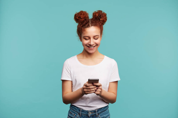Positive young pretty curly female wearing her foxy hair while in knot posing over blue background with mobile phone in raised hands and looking cheerfully on screen - Photo, Image