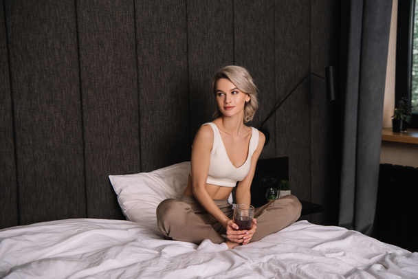 atraente jovem segurando copo de vinho tinto enquanto sentado na cama com pernas cruzadas
 - Foto, Imagem