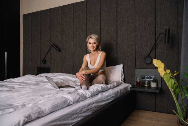 houkutteleva nainen katselee kameraa istuessaan sängyssä lähellä kannettavaa tietokonetta
 - Valokuva, kuva