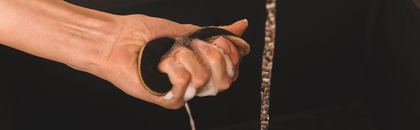 καλλιεργημένη όψη θηλυκού χεριού με υγρό σφουγγάρι σαπουνιού κοντά στο νερό που απομονώνεται σε μαύρη, οριζόντια εικόνα - Φωτογραφία, εικόνα