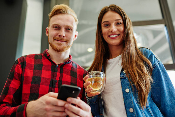 Portrait of cheerful man and woman looking at camera while enjoying fresh beverage and communication via cellphone, positive millennial users waiting for message about discounts in web store - Foto, Imagen