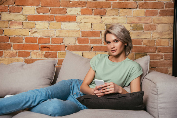 attractive, confident woman sitting of sofa, chatting on smartphone and looking at camera - Zdjęcie, obraz