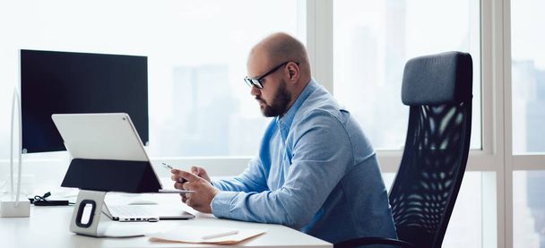 Vista lateral del hombre calvo adulto en gafas y camisa azul sentado en el escritorio con aparatos modernos en la oficina y el uso de teléfonos inteligentes
 - Foto, imagen