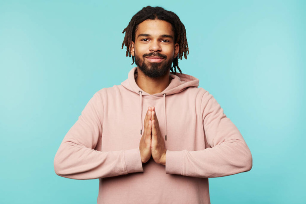 Positive young brown-eyed brunette male with dreadlocks smiling pleasantly at camera and raising hands in praying gesture, standing over blue background - Фото, зображення