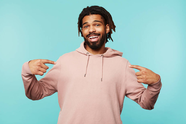 Glad young brown-eyed brunette male with dreadlocks smiling cheerfully at camera while pointing on himself with raised forefingers, standing over blue background - Foto, afbeelding