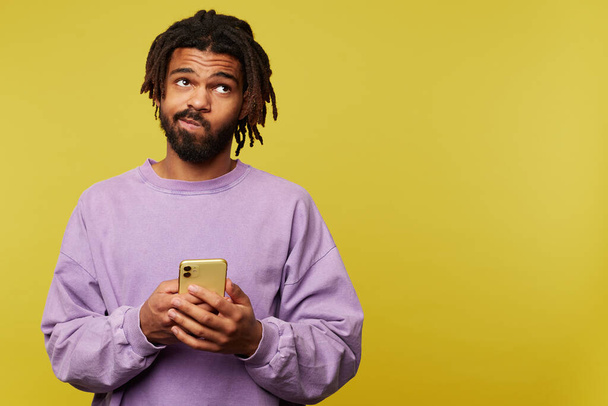 Puzzled young handsome brunette bearded male with dark skin twisting his mouth while looking pensively upwards, standing over yellow background with smartphone - Valokuva, kuva
