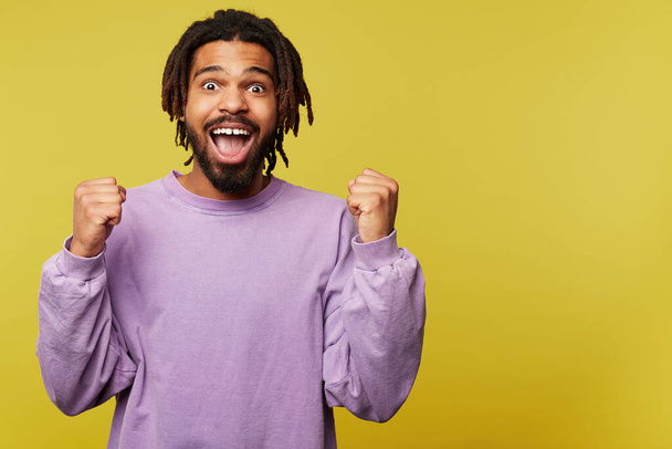 Excited young lovely dark skinned brunette man with dreadlocks looking surprisedly at camera with opened mouth and keeping his hands raised, isolated over yellow background - Фото, зображення