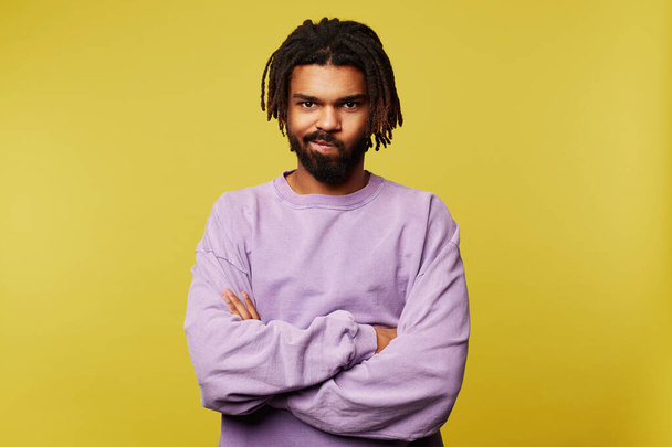 Offended young pretty dark skinned brunette male in purple sweatshirt crossing arms on chest and twisting mouth while looking at camera, isolated over yellow background - Fotoğraf, Görsel