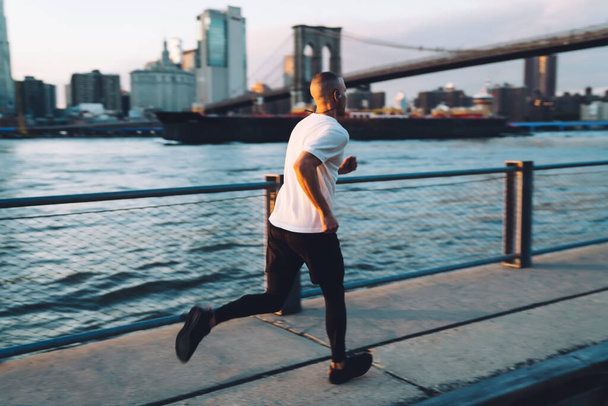 Young male in sportswear and earphones jogging fast while looking straight in morning on urban background with bridge and ship. Motion blurr velocity effect - Foto, imagen