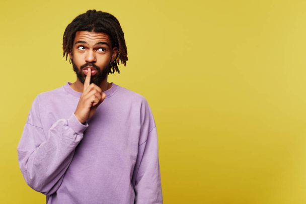 Studio photo of young attractive dark skinned bearded man keeping forefinger on his mouth while asking to keep secret, isolated over yellow background - Fotografie, Obrázek
