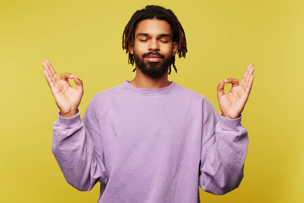 Pleasant looking young dark skinned bearded male in purple sweatshirt keeping his eye closed while standing over yellow background and raising hands with mudra gesture - Fotografie, Obrázek