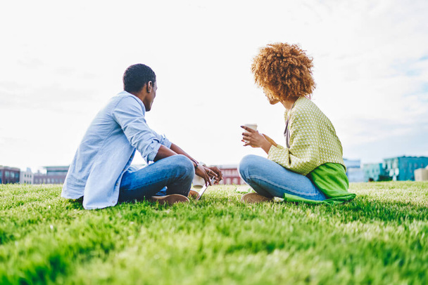 Back view of two african american hipster friends dressed in casual wear sitting on green grass in park.Couple in love enjoying coffee beverage and resting in urban setting in sunny day - Foto, afbeelding