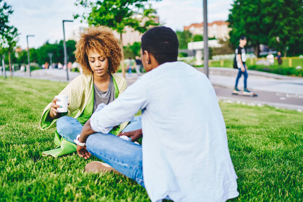 Two dark skinned friends talking with each other and spending leisure time together in urban setting in green park.African american girlfriend communicating with boyfriend drinking tasty coffee - 写真・画像