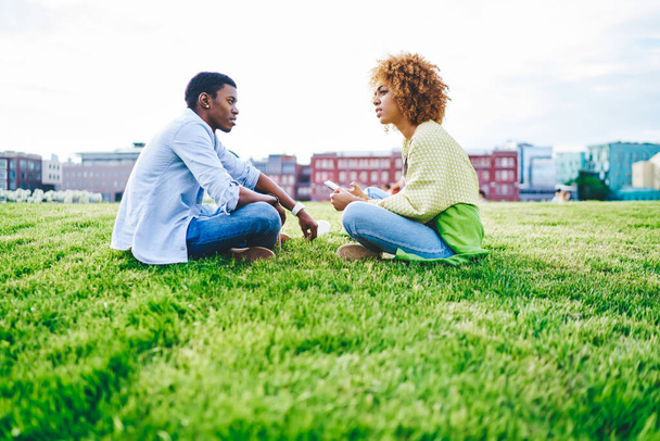 Best african american friends dressed in casual wear resting in park sitting in lotus pose on green grass and communicating with each other.Dark skinned female with smartphone talking with boyfriend - Φωτογραφία, εικόνα
