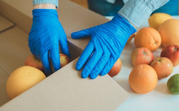 Volunteer in protective medical gloves putting fruits In donation box. - Photo, Image