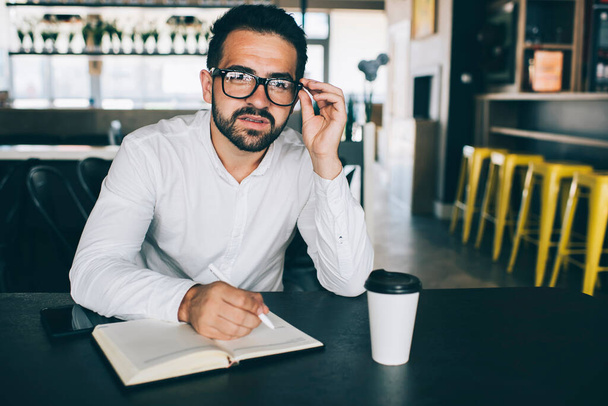 Portrait of serious male entrepreneur with personal planner sitting at cafeteria table and looking at camera, Caucasian bearded man in classic glasses for vision protection working with trade strategy - 写真・画像