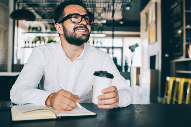 Successful man in optical spectacles spending time for planning productive strategy with personal textbook for memo notes, happy male with coffee to go pondering on checklist for startup project - Foto, imagen