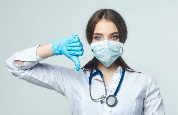 Covid19, coronavirus, a concept of healthcare and doctors. Portrait of a female doctor, shows that the bad result is a thumbs down, puts on a medical mask to prevent infection with the virus, white coat - Photo, image