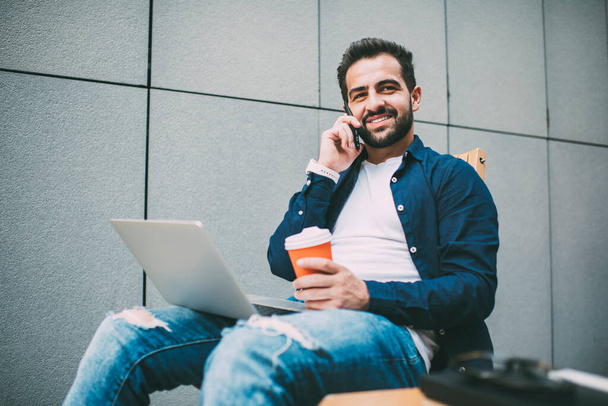 Happy millennial man in casual apparel calling to friend for communicate during coffee break from online programming via netbook, cheerful male generation using roaming connection on cellular - Zdjęcie, obraz