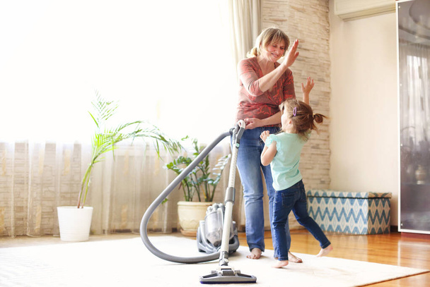 Full length happy mature woman with vacuum cleaner giving high five to little girl while tidying cozy living room at home together - Fotoğraf, Görsel