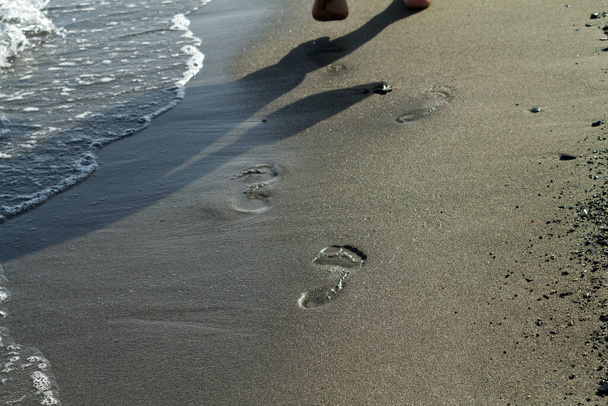 Spiaggia di sabbia con un'onda di mare schiumosa, impronte e un'ombra umana alla luce del sole
 - Foto, immagini