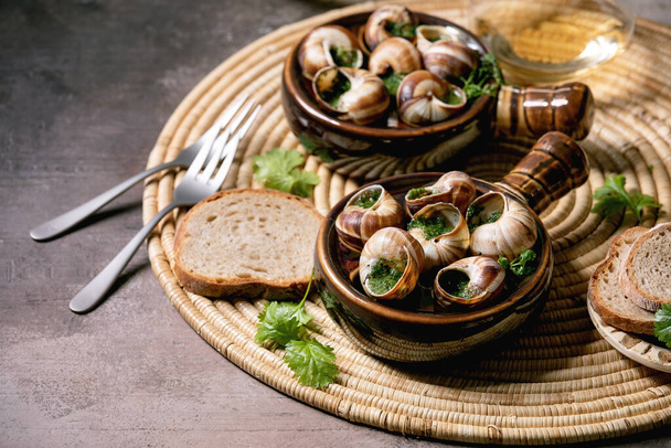 Escargots de Bourgogne - Snails with herbs butter, gourmet dish, in two traditional ceramic pans with coriander, bread and glass of white wine on straw napkin over brown textured background. - Fotografie, Obrázek