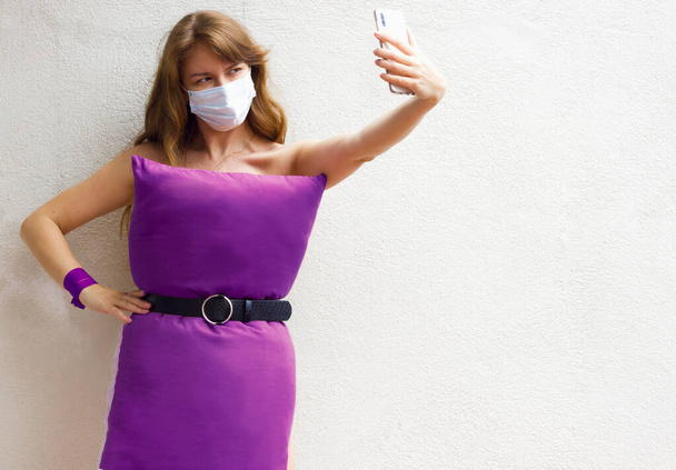 Γυναίκα σε ιατρική μάσκα, μωβ φόρεμα μαξιλάρι λήψη selfie φωτογραφία στο κινητό τηλέφωνο για τρελό πρόκληση μαξιλάρι οφείλεται να μείνετε στο σπίτι απομόνωση σε λευκό φόντο τοίχο. Κορίτσι της μόδας. Καροναβιάρι Καραντίνα. - Φωτογραφία, εικόνα