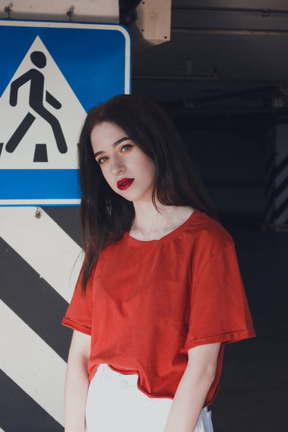 Mujer joven caminando en la ciudad. Retrato de una chica elegante y de moda estacionada cerca de una señal. Chica en una camiseta roja y pantalones blancos. - Foto, Imagen