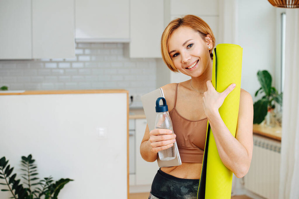 Happy shiny woman smiling after yoga practice, looking at camera. She's holding mat and water bottle. She's home in her kitchen. - Фото, изображение