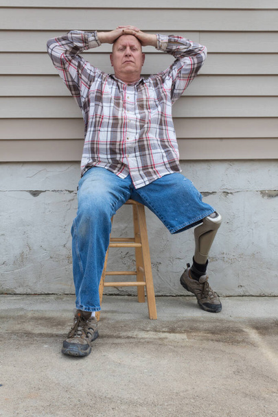 Homem amputado sentado em um banco, prótese perna para fora, mãos na cabeça olhando para a câmera, espaço de cópia, aspecto vertical
 - Foto, Imagem